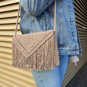May include: A beige macrame shoulder bag with a woven pattern and fringe detail. The bag has a long strap and a flap closure. The bag is made of a natural fiber material and is a neutral color.