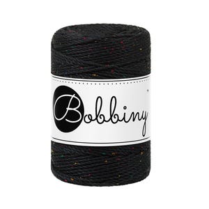 Puede incluir: Un carrete de hilo de macramé negro con motas multicolores. El hilo está enrollado alrededor de un núcleo y tiene una etiqueta blanca con la palabra "Bobbiny" en escritura cursiva.