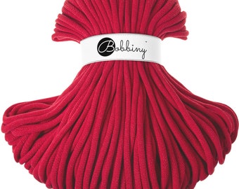 May include: A vibrant red spool of Bobbiny macrame cord. The cord is tightly wound and held together with a white band featuring the Bobbiny logo. The texture of the cord is visible, suggesting a soft, yet durable material, perfect for crafting projects.