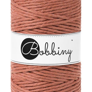 Puede incluir: Un carrete de hilo de macramé de color terracota. El hilo está bien enrollado y envuelto con una etiqueta blanca con el nombre de la marca "Bobbiny" en escritura negra estilizada. El hilo parece estar hecho de un material suave y tejido.
