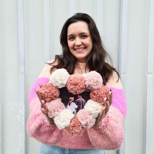 May include: A pink and white heart-shaped wreath made of pom-poms with a photo inside. The wreath is being held by a person wearing a pink and white striped sweater.