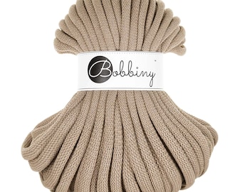 May include: A beige cotton cord, wound into a circle, with a white label that reads "Bobbiny" in black lettering.