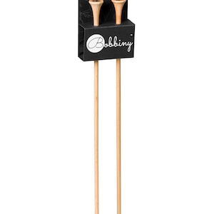 May include: A pair of wooden knitting needles with a black cardboard holder. The holder features the word "Bobbing" in a cursive font. The needles are light brown and have a pointed tip. The holder has a small hole at the top.