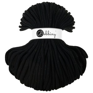 Puede incluir: Un ovillo de cordón de macramé negro con una etiqueta blanca que dice "Bobbing" en cursiva. El cordón está bien enrollado, mostrando una superficie texturizada con motas de color. Ideal para manualidades.