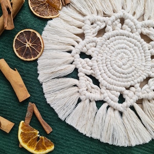 Peut inclure: Un dessous de verre en crochet blanc avec une bordure en franges. Le dessous de verre a un design circulaire avec un cercle central et un motif de nœuds qui l'entoure.