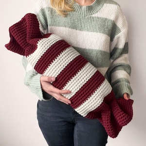 May include: A burgundy and cream striped, crocheted hot water bottle cover. The cover is cylindrical with a ruffled end. The person holding the cover is wearing a green and white striped sweater and blue jeans.