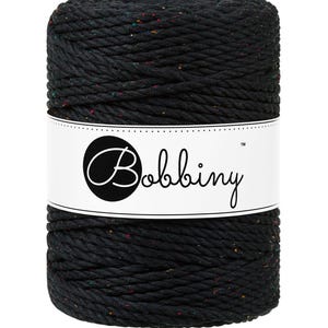 Puede incluir: Un carrete cilíndrico de cordón de macramé negro con motas de color. Una etiqueta blanca con la palabra "Bobbiny" en cursiva envuelve el centro. El cordón parece estar bien enrollado, adecuado para varios proyectos de manualidades.