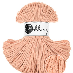 Może przedstawiać: Szpula brzoskwiniowego sznurka do makramy Bobbiny. Sznurek jest ciasno nawinięty i posiada szczegółowy widok z bliska na tkaną teksturę. Etykieta na szpuli wyświetla nazwę marki "Bobbiny" w stylowym skrypcie.