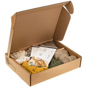 Puede incluir: Una caja de cartón abierta que contiene suministros para macramé. La caja contiene varios colores de cuerda de algodón gruesa, incluyendo crema, amarillo mostaza, verde oliva y gris. También se incluyen instrucciones y clavijas de madera.