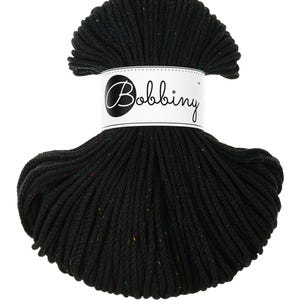 Puede incluir: Un gran ovillo de hilo negro, bien enrollado, con motas de color. Una etiqueta blanca con la palabra "Bobbing" en escritura cursiva está envuelta alrededor del centro del hilo. El hilo parece ser un cordón grueso y texturizado.