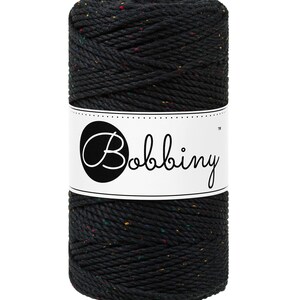 Puede incluir: Un carrete negro de cordón de macramé de la marca Bobbin. El cordón tiene motas de color. Una etiqueta blanca con la palabra "Bobbin" en escritura cursiva envuelve el centro del carrete. Listo para proyectos de manualidades.