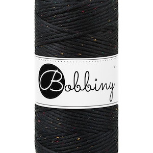 Puede incluir: Un carrete cilíndrico de cordón de macramé negro con motas de colores. El cordón está enrollado sobre sí mismo, y una etiqueta blanca con la palabra "Bobbiny" en cursiva está envuelta alrededor del centro.