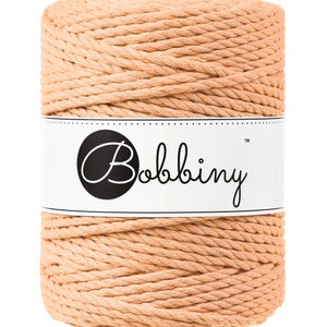 May include: A cylindrical spool of peach-colored macrame cord. A white label encircles the spool, displaying the brand name "Bobbiny" in a black, elegant script. The cord is tightly wound, showcasing a textured, woven appearance.