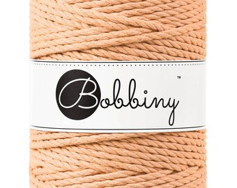 May include: A cylindrical spool of peach-colored macrame cord. A white label encircles the spool, displaying the brand name "Bobbiny" in a black, elegant script. The cord is tightly wound, showcasing a textured, woven appearance.