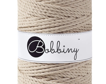 Pode incluir: Um carretel de corda de algodão bege com o nome da marca "Bobbiny" impresso em um rótulo branco.