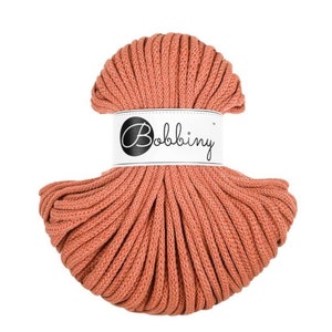 Puede incluir: Una madeja de cordón de macramé Bobbiny de color coral. El cordón está bien enrollado y tiene un aspecto texturizado. Una etiqueta blanca con el nombre de la marca "Bobbiny" está envuelta alrededor del centro de la madeja. Ideal para proyectos de manualidades.