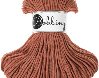 Könnte beinhalten: Eine Spule aus terracottafarbenem Baumwollgarn, spiralförmig gewickelt. Das Garn ist mit "Bobbiny" in schwarzer Schrift auf einem weißen Etikett beschriftet.