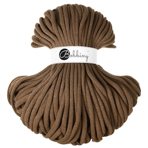Pode incluir: Um novelo de fio de macramé castanho. Uma faixa branca com a palavra "Bobbiny" em fonte cursiva envolve o centro. O fio é de uma cor castanha terrosa, adequado para vários projetos de artesanato e decoração.