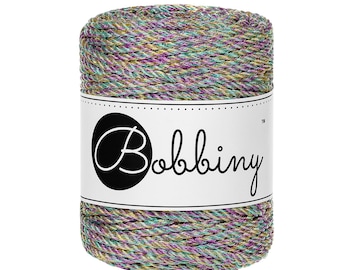 May include: A cylindrical spool of braided cord featuring a multi-colored metallic finish with shimmering green, purple, and gold threads. A white paper label wraps around the center, displaying the brand name Bobbiny in black script text.