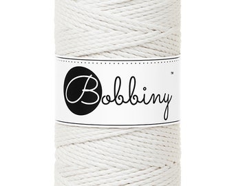 Könnte beinhalten: Eine Spule weißes Makramee-Garn mit dem Markennamen "Bobbiny" auf einem Etikett. Das weiße Etikett hat schwarzen Text und einen schwarzen Kreis. Das Garn ist eng gewickelt und eignet sich für verschiedene Bastel- und DIY-Projekte. Das Produkt ist zum Basteln.