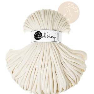 Può includere: Un grande rotolo di corda di cotone color crema chiaro etichettato "Bobbiny Jumbo Soft 10mm Braided Cord". L'etichetta è bianca con testo nero. Un piccolo cerchio con un'icona di piuma e la parola "SOFT" si trova nell'angolo in alto a destra.
