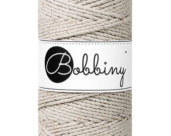 May include: A cylindrical spool of beige macrame cord. The cord is tightly wound and has small flecks of color throughout. A white label with the word "Bobbiny" in a script font is wrapped around the center of the spool.