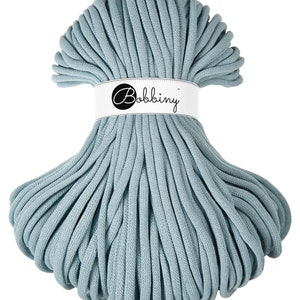 Puede incluir: Un ovillo de cordón de macramé azul claro. El cordón está envuelto con una etiqueta blanca que dice "Bobbiny" en una fuente negra. El cordón está bien enrollado y listo para proyectos de manualidades.
