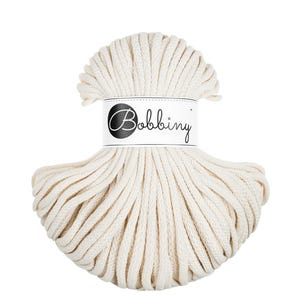 Puede incluir: Cordón de macramé color crema, enrollado firmemente en un ovillo. Una etiqueta blanca con la palabra "Bobbing" en fuente cursiva está envuelta alrededor del centro. El cordón es adecuado para varios proyectos de manualidades.