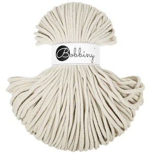 May include: A bundle of off-white macrame cord. The cord is tightly wound and secured with a white label that reads "Bobbiny" in a cursive font. The cord is a neutral color, suitable for various crafting projects.
