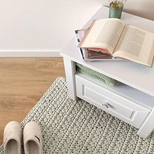 May include: A domestic scene with a white side table holding an open book, a small vase with dried flowers, and magazines. A light green knitted rug and a pair of beige slippers are on the wooden floor.