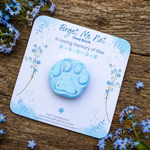 May include: A light blue seed bomb shaped like a paw print, on a card with the text "Forget Me Not" and "In loving memory of Max." The card features blue floral illustrations and hearts, with instructions.