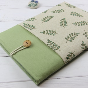 Puede incluir: Funda de tela verde claro y beige con cierre de botón. La sección beige presenta un patrón repetido de hojas verdes. La funda está diseñada para guardar una tableta o un libro. El botón es de madera y está sujeto con una cuerda blanca.