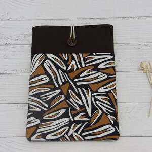 Puede incluir: Una funda para portátil con estampado marrón y blanco con cierre de botón marrón. La funda está hecha de tela y tiene una solapa marrón en la parte superior.