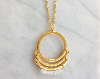May include: A gold necklace with a geometric pendant featuring a double circle design and a row of white stones.