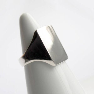 May include: A silver signet ring with a square face. The ring is displayed on a white cone-shaped ring holder.