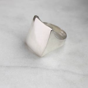 May include: A silver ring with a large, flat, square-shaped top. The ring is made of smooth, polished metal.