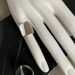 May include: Two silver rings, one square and one rounded, on a mannequin hand. The rings are both simple and modern in design.