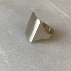 Pode incluir: Um anel de prata com uma parte superior retangular grande e plana. A parte superior do anel &eacute; ligeiramente curva e tem um acabamento escovado.