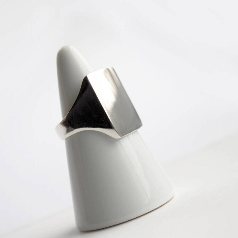 May include: A silver signet ring with a square face. The ring is displayed on a white cone-shaped ring holder.