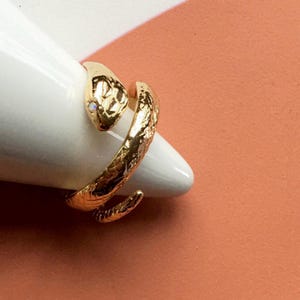 Puede incluir: Un anillo de serpiente dorado con un ojo de piedra transparente. El anillo está enrollado y tiene una superficie texturizada.