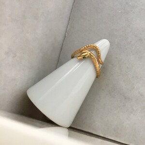 May include: Gold snake ring with a green stone eye. The ring is on a white ring holder.