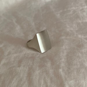 Pode incluir: Um anel de sinete de prata com acabamento escovado e uma face quadrada.