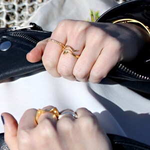 May include: A close-up of a person's hands wearing several gold rings. The rings are stacked on the fingers and feature a variety of designs, including a snake-like ring and a ring with a textured band.