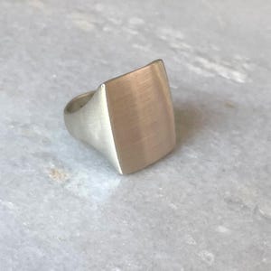 May include: A silver ring with a square shaped top. The top of the ring has a brushed finish.