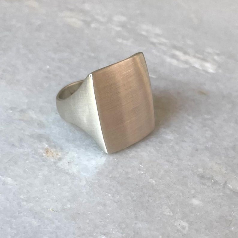 May include: A silver ring with a square shaped top. The top of the ring has a brushed finish.