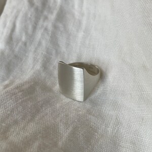 Pode incluir: Um anel de prata com acabamento escovado e topo quadrado. O anel est&aacute; sobre um fundo de tecido branco.