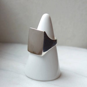 May include: A silver signet ring with a square top. The ring is displayed on a white ring holder.