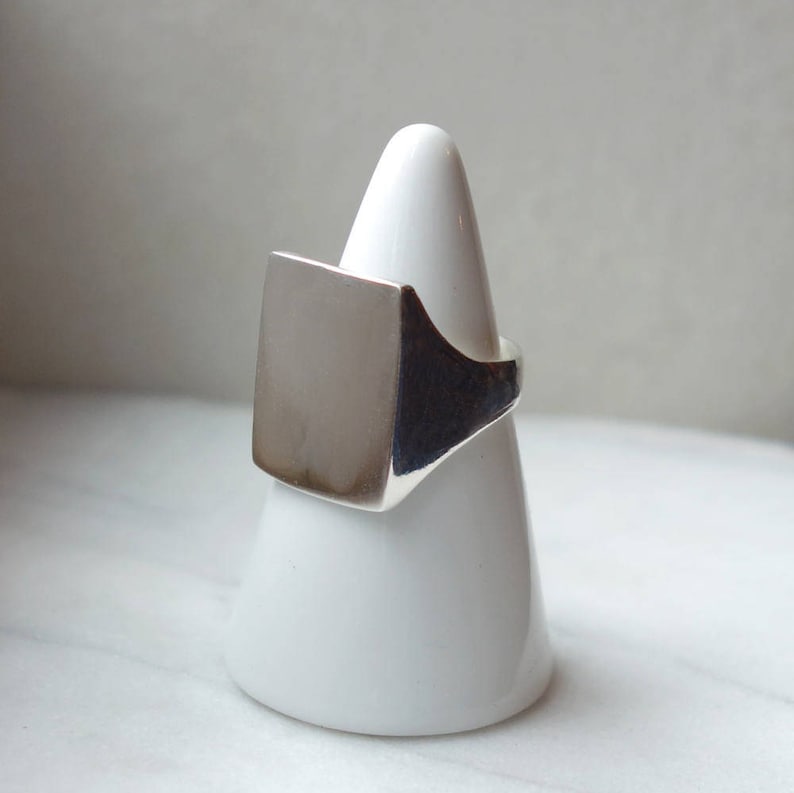 May include: A silver signet ring with a square top. The ring is displayed on a white ring holder.