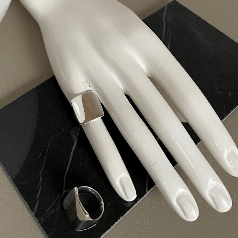 May include: Two silver rings, one square and one rounded, on a mannequin hand. The rings are both simple and modern in design.