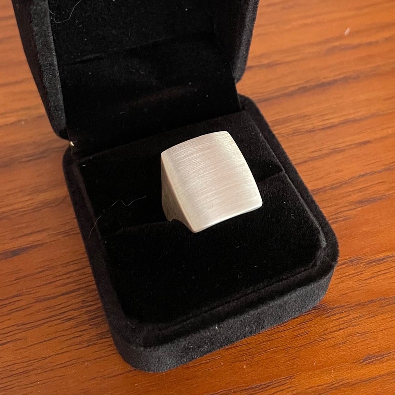 May include: A silver square ring with a brushed finish, displayed in a black velvet ring box.